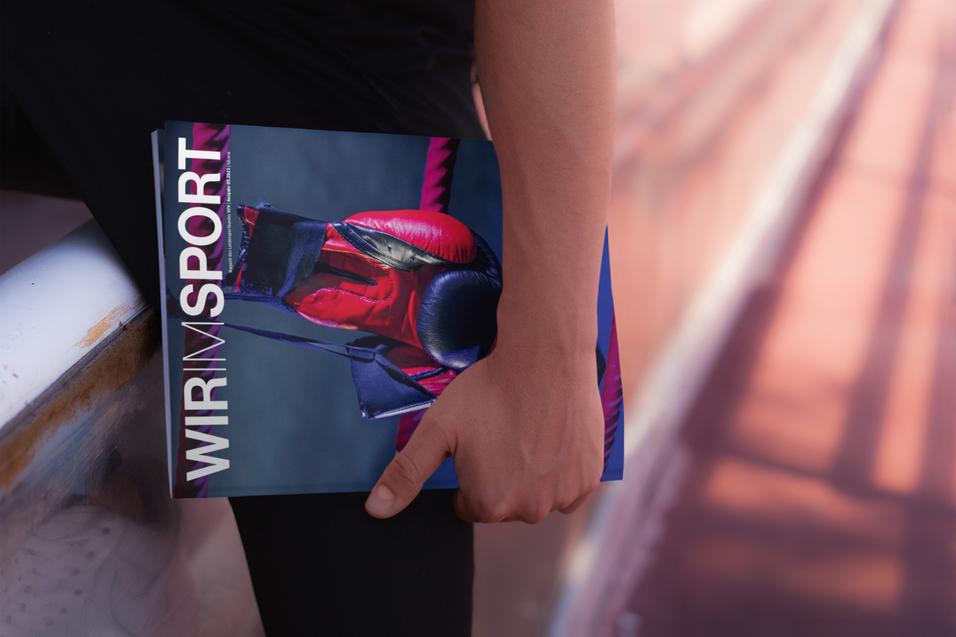 Hand hält eine Zeitschrift mit dem Titel „Wir im Sport“ in lebhaften Farben, auf einer Sportfläche platziert.